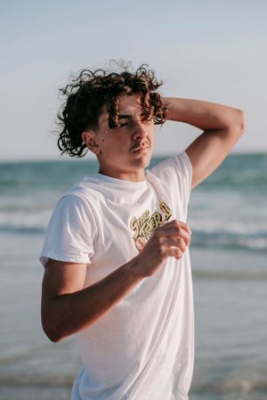 Person in white t-shirt poses expressively against ocean backdrop.