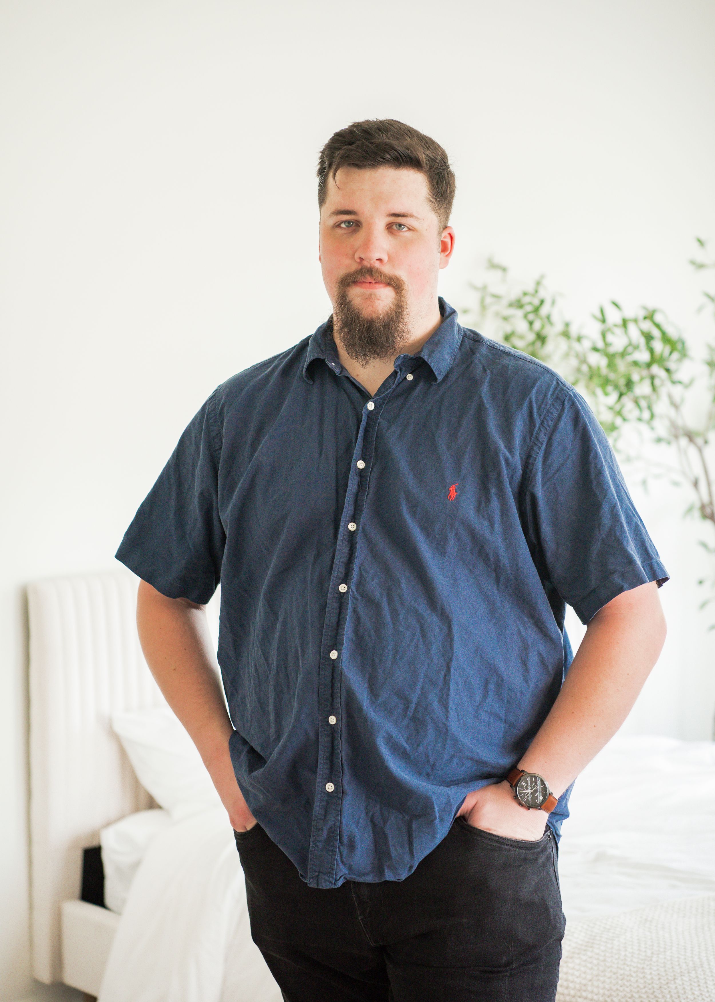 Man in navy button-down shirt stands casually against white wall interior.