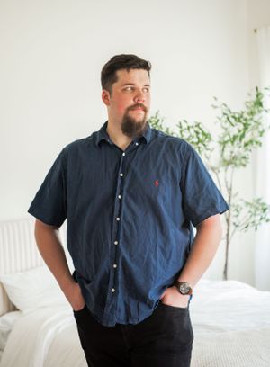 Casual portrait of man in blue shirt standing against white wall with hands in pockets.