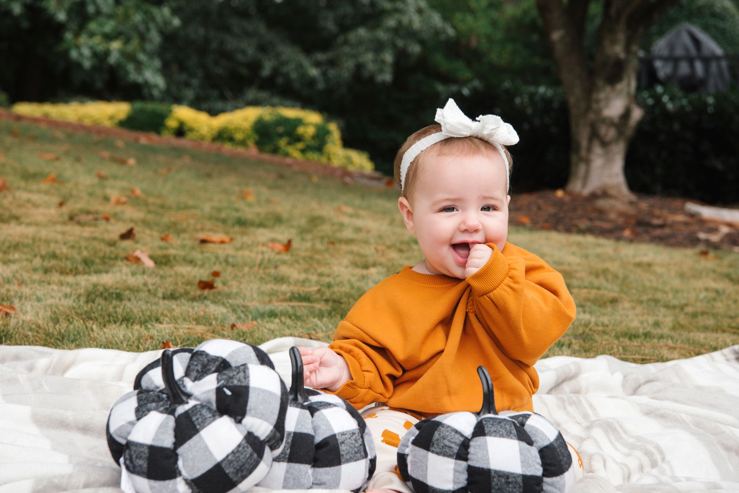 Baby in orange sweater sits on buffalo plaid blanket outdoors with white bow headband.