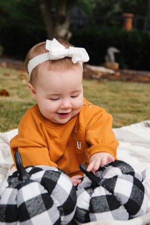 Smiling baby wearing orange sweater and white bow plays with black and white checkered pillows.