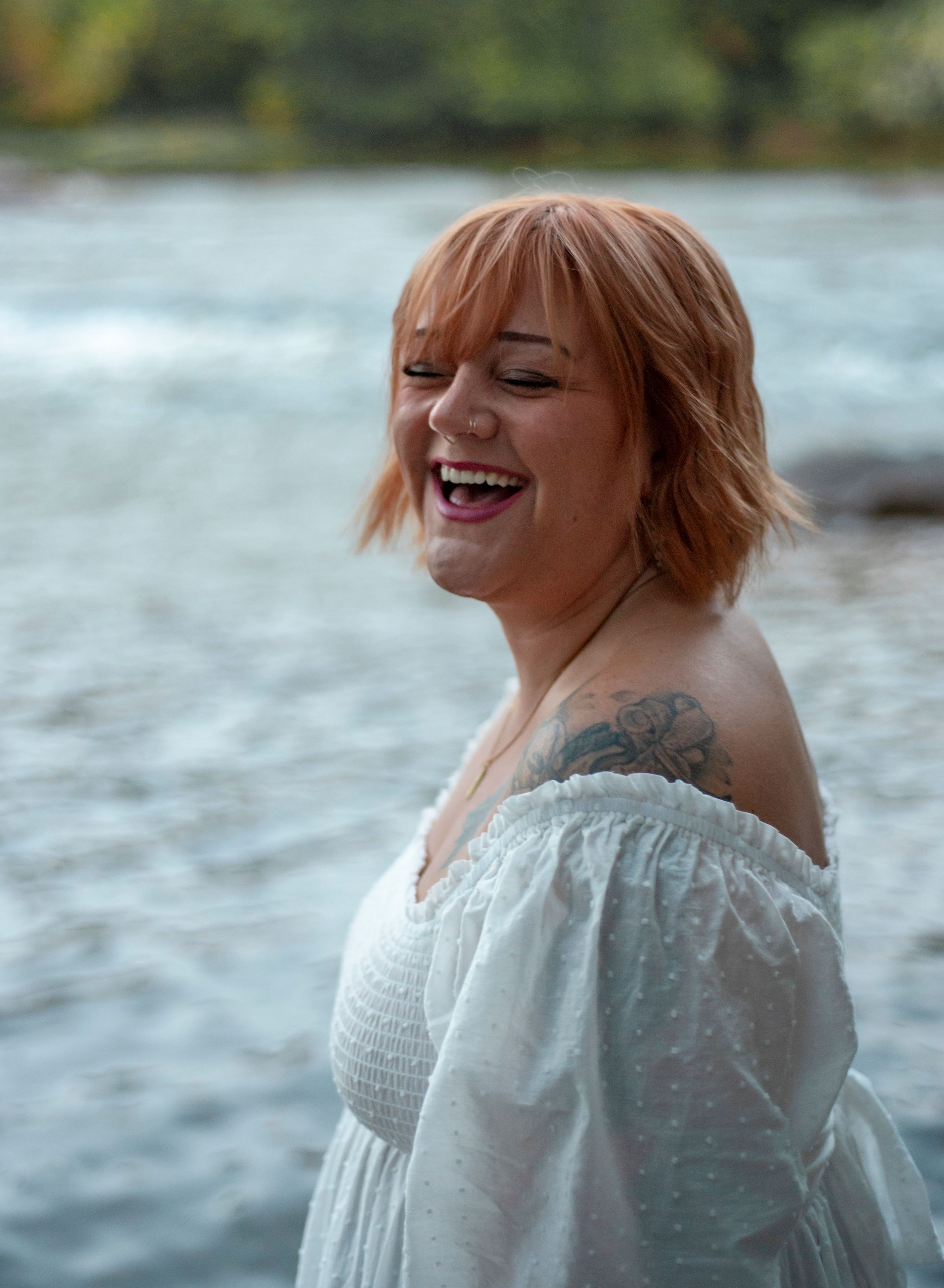 Joyful portrait by river wearing white off-shoulder dress with short hair.