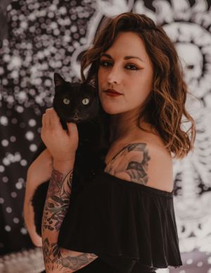 Portrait of a tattooed woman in black holding a dark cat against a sparkly background.