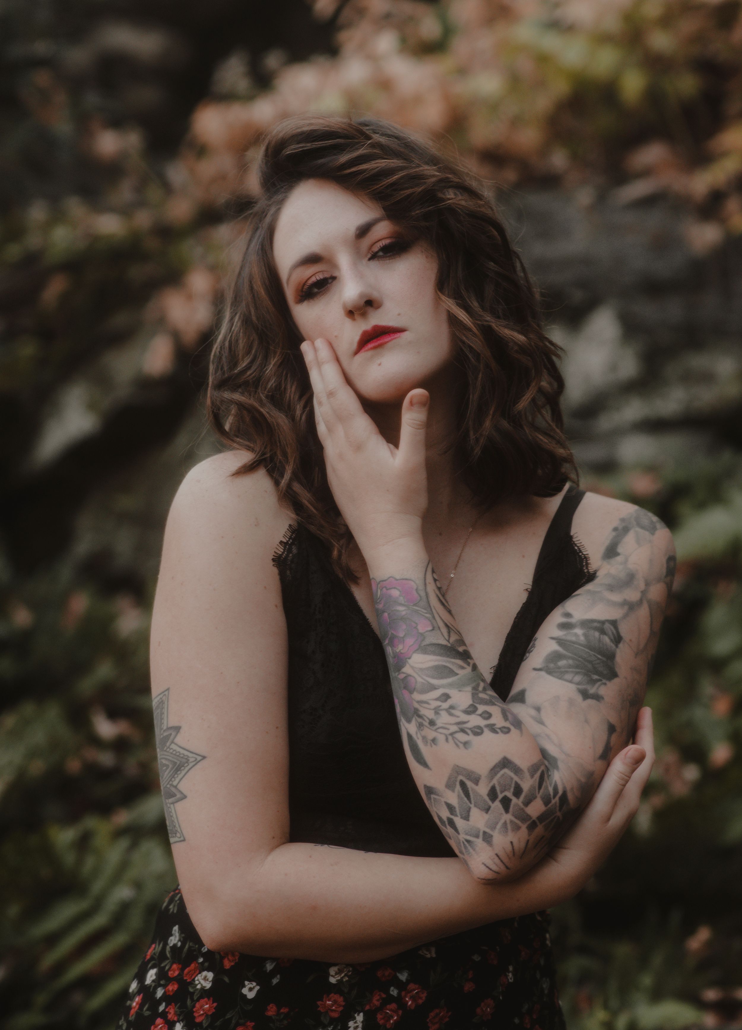 Moody portrait of a tattooed woman in black against an autumn forest backdrop.