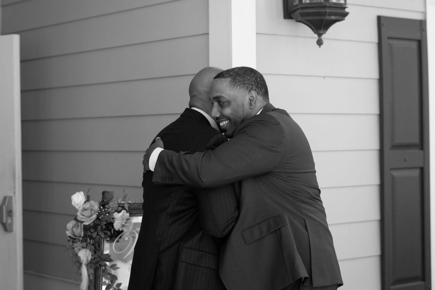 Two people share an emotional embrace during a wedding ceremony outside a white house.