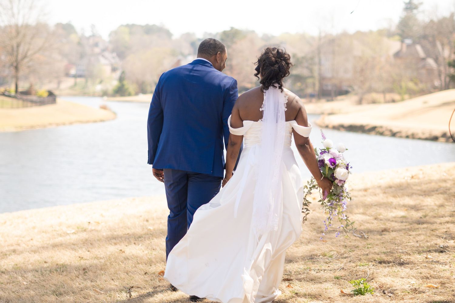 Newlyweds walking together along a lakeside path with the bride holding a bouquet.