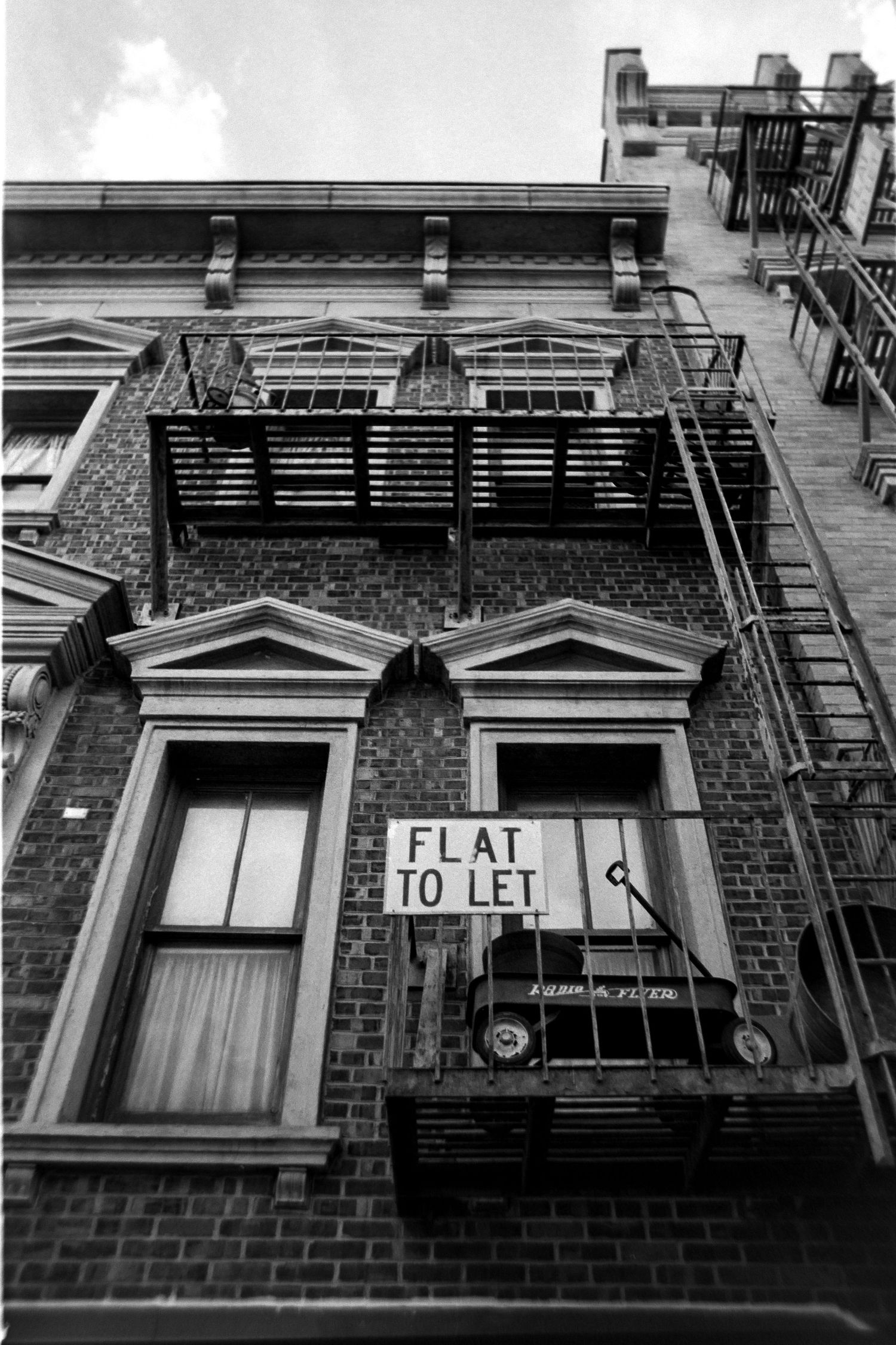 Black and white photo of a brick building facade with two arched windows and a 'FLAT TO LET' sign mounted between them.