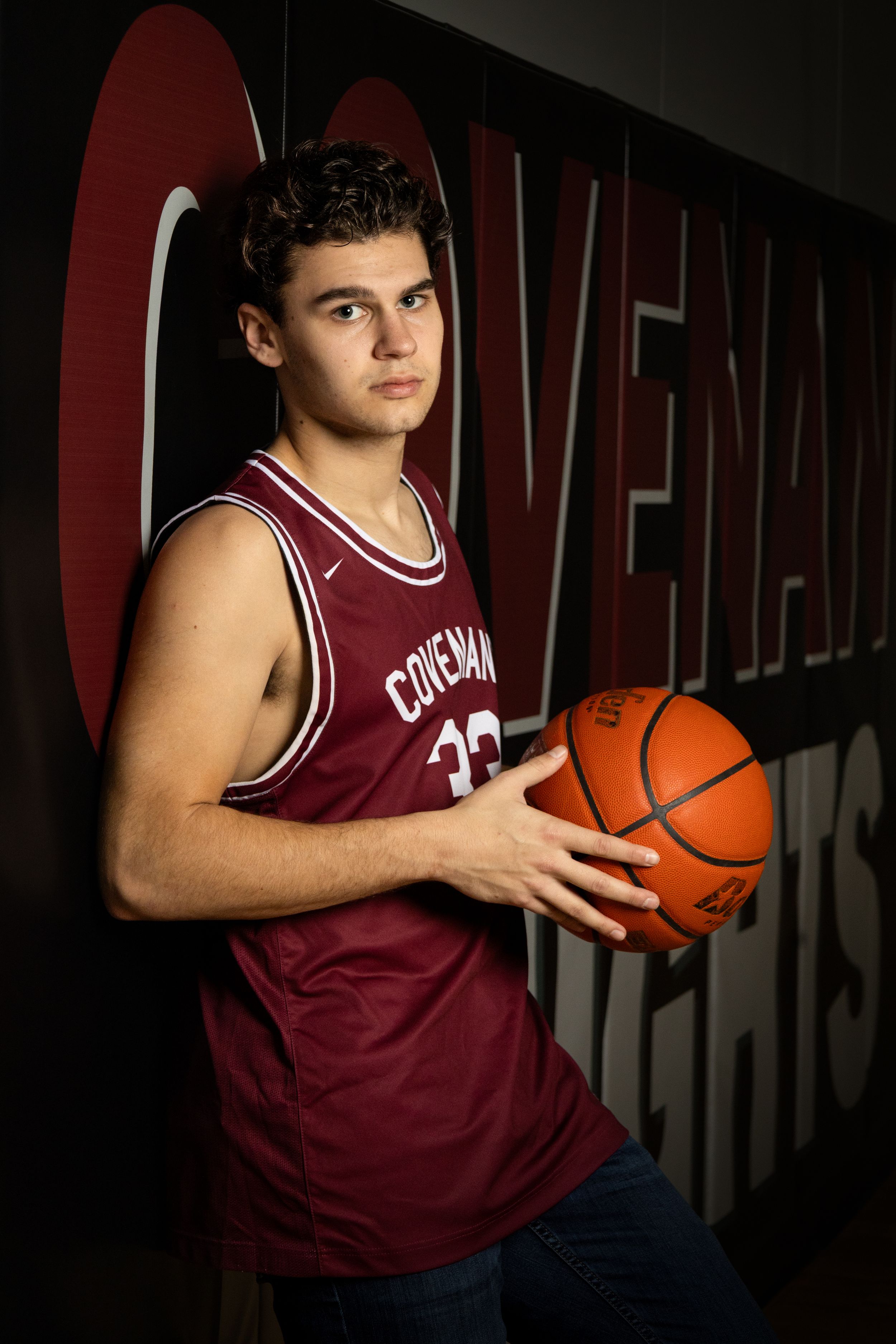 A basketball player in a maroon Cincinnati jersey poses with a basketball against a dark background.