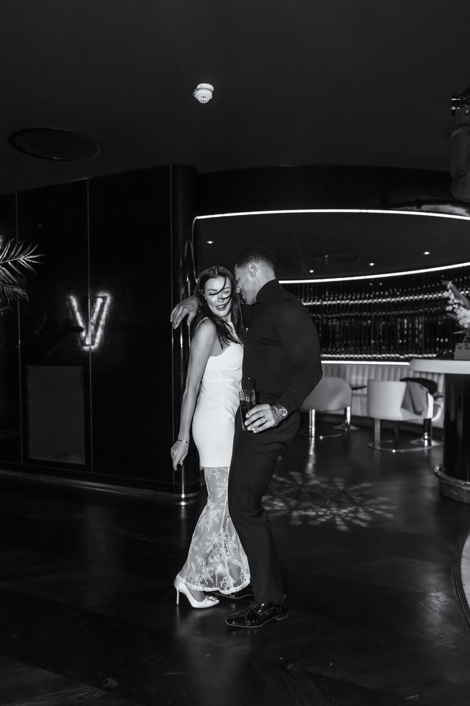 A couple shares an intimate dance moment in black and white on a dimly lit dance floor with a lace wedding dress.