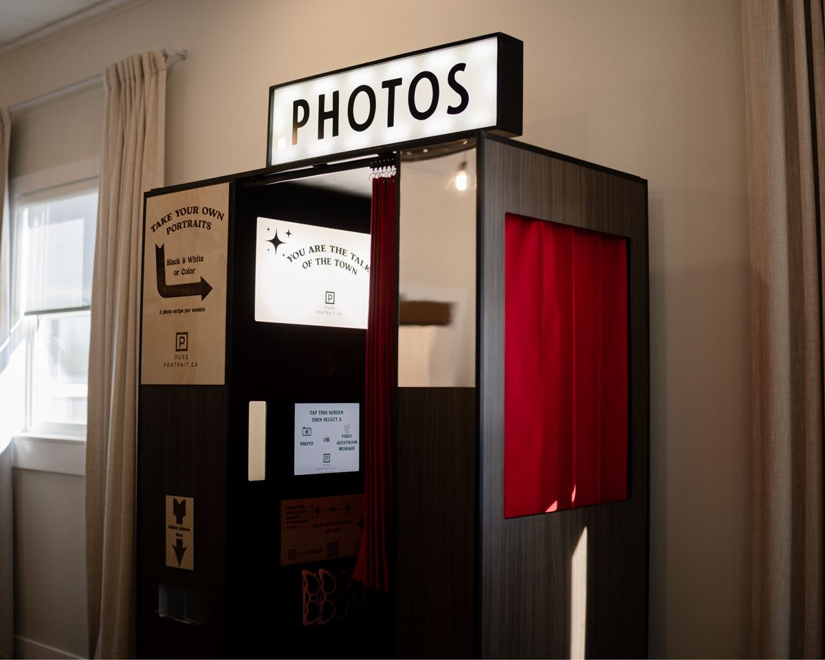 A retro photo booth with a red curtain and illuminated 'PHOTOS' sign mounted on top stands against a beige wall.
