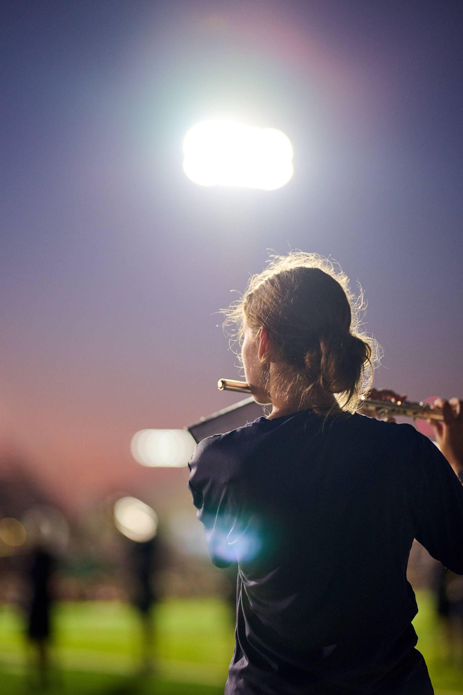 Flutist silhouetted against evening sky with stadium lighting creating dramatic backlighting.