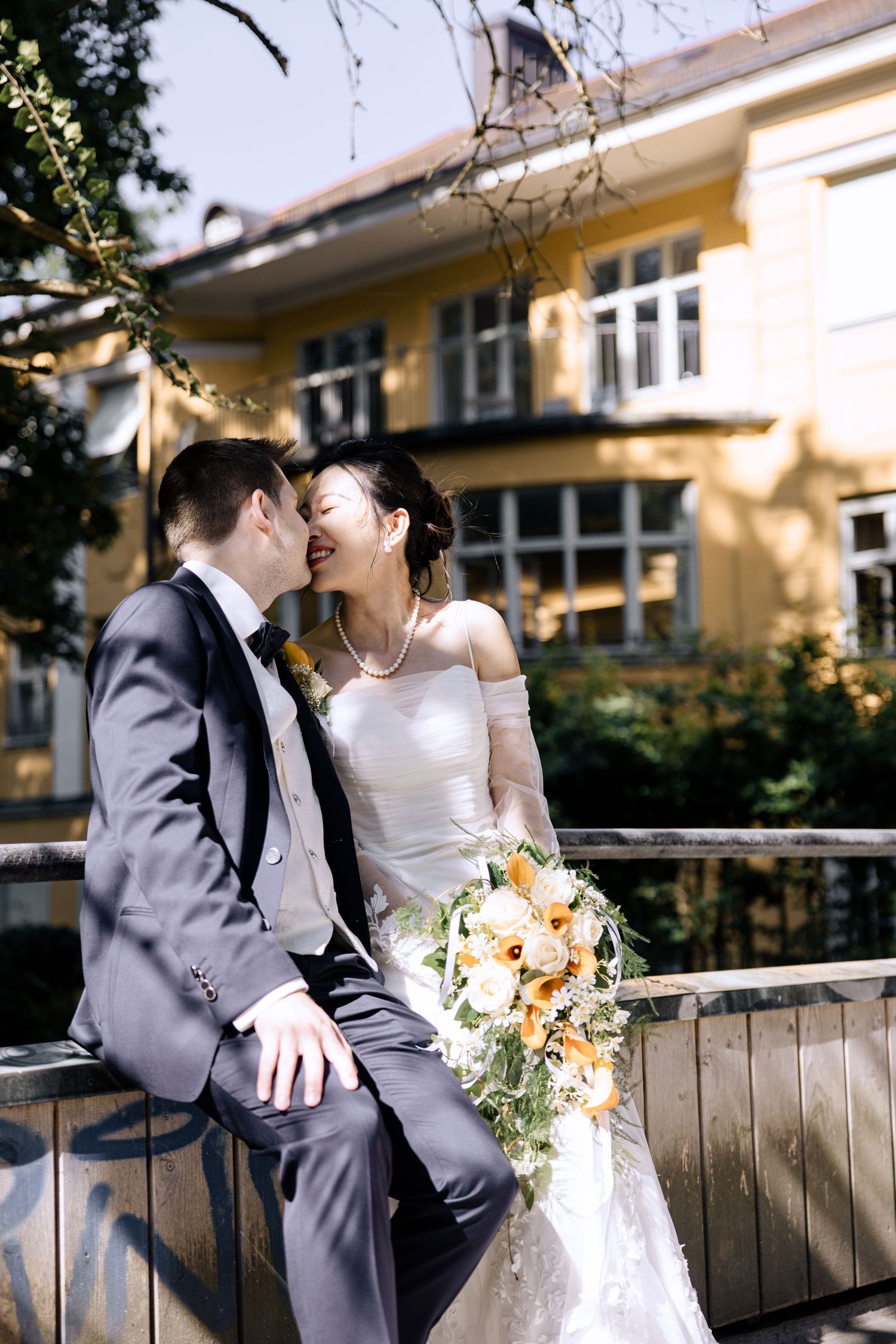 Wedding couple sharing intimate moment on wooden bench outside yellow building.