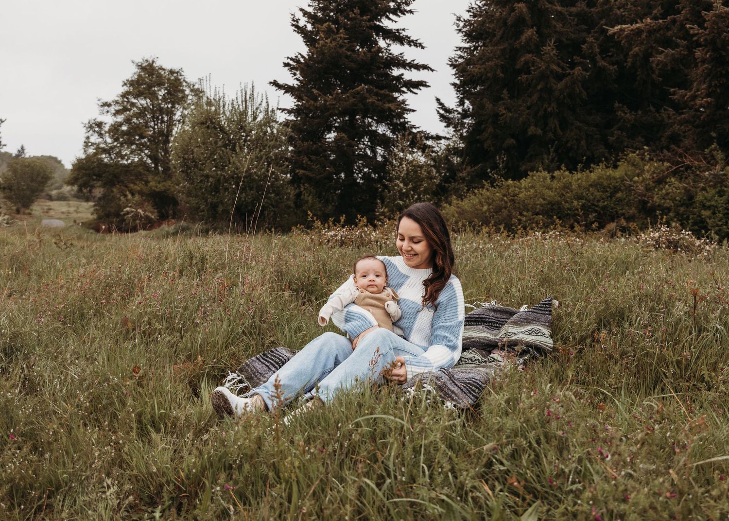 A cozy autumn scene with people relaxed on a blanket in a grassy field surrounded by evergreen trees.