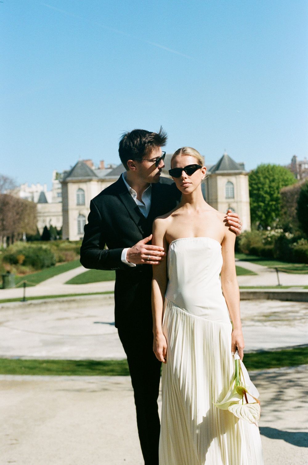 Elegant couple in formal attire poses outdoors with stately buildings and gardens in the background on a sunny day.