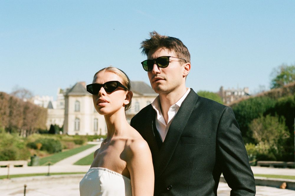 Two people in sunglasses pose elegantly outdoors against a backdrop of manicured gardens and classical architecture.