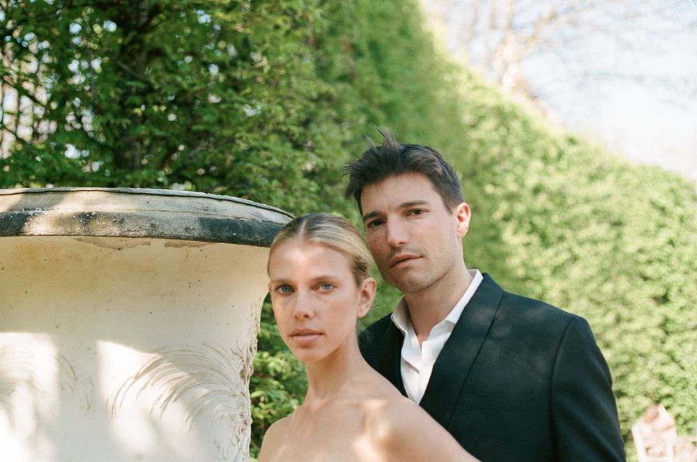An elegant couple poses outdoors next to a decorative stone urn in a formal garden setting.