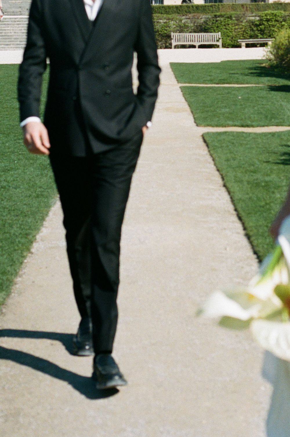 A person in a black suit and dress shoes walks on a concrete path bordered by manicured green lawns and a garden bench.