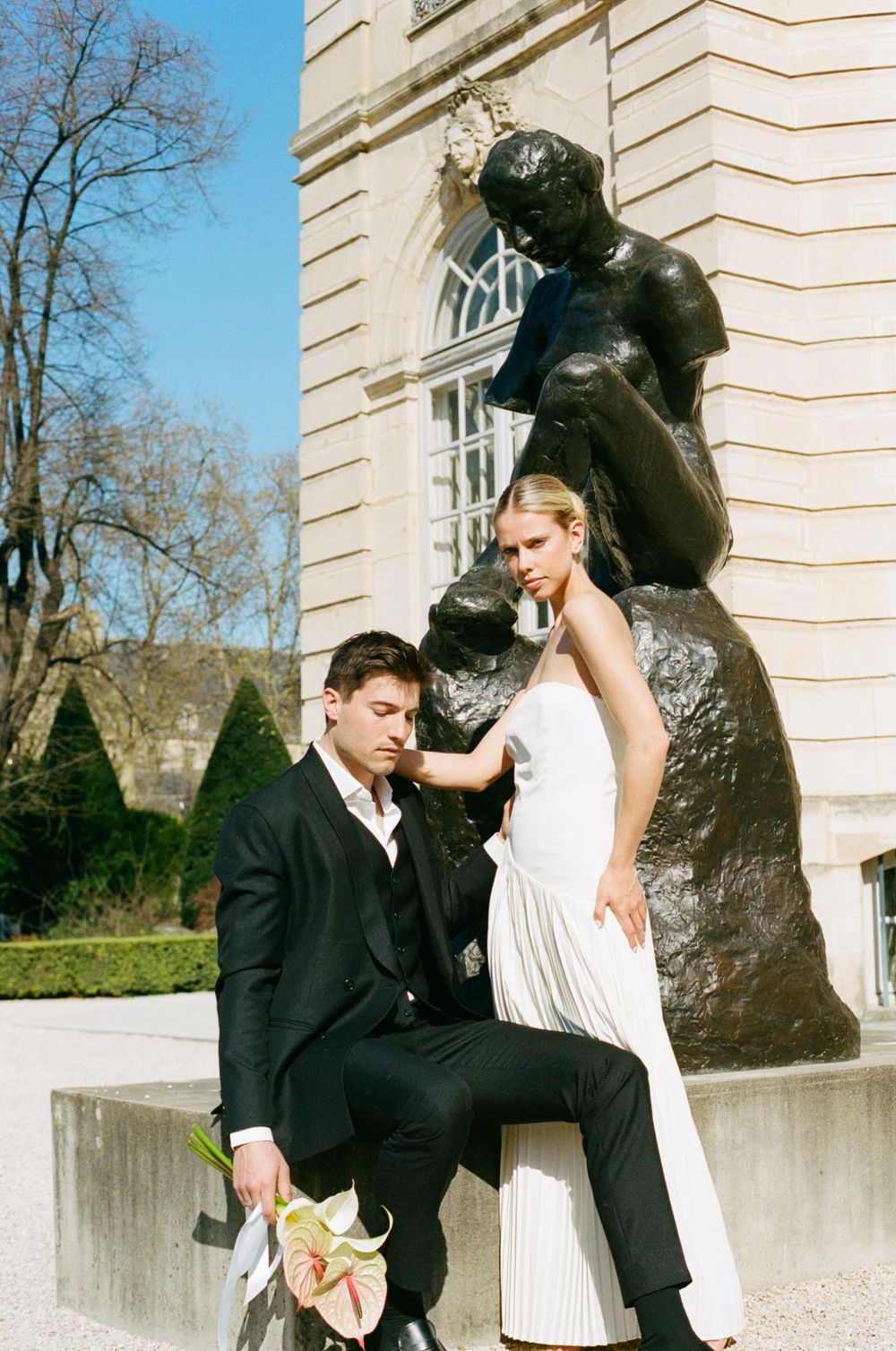 Elegant couple poses beside large bronze sculpture outside historic building with arched windows.