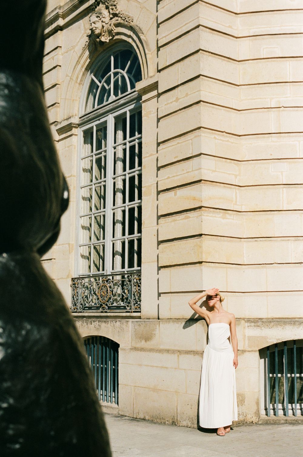 Person in flowing white dress poses against ornate stone building with tall arched window and decorative ironwork.