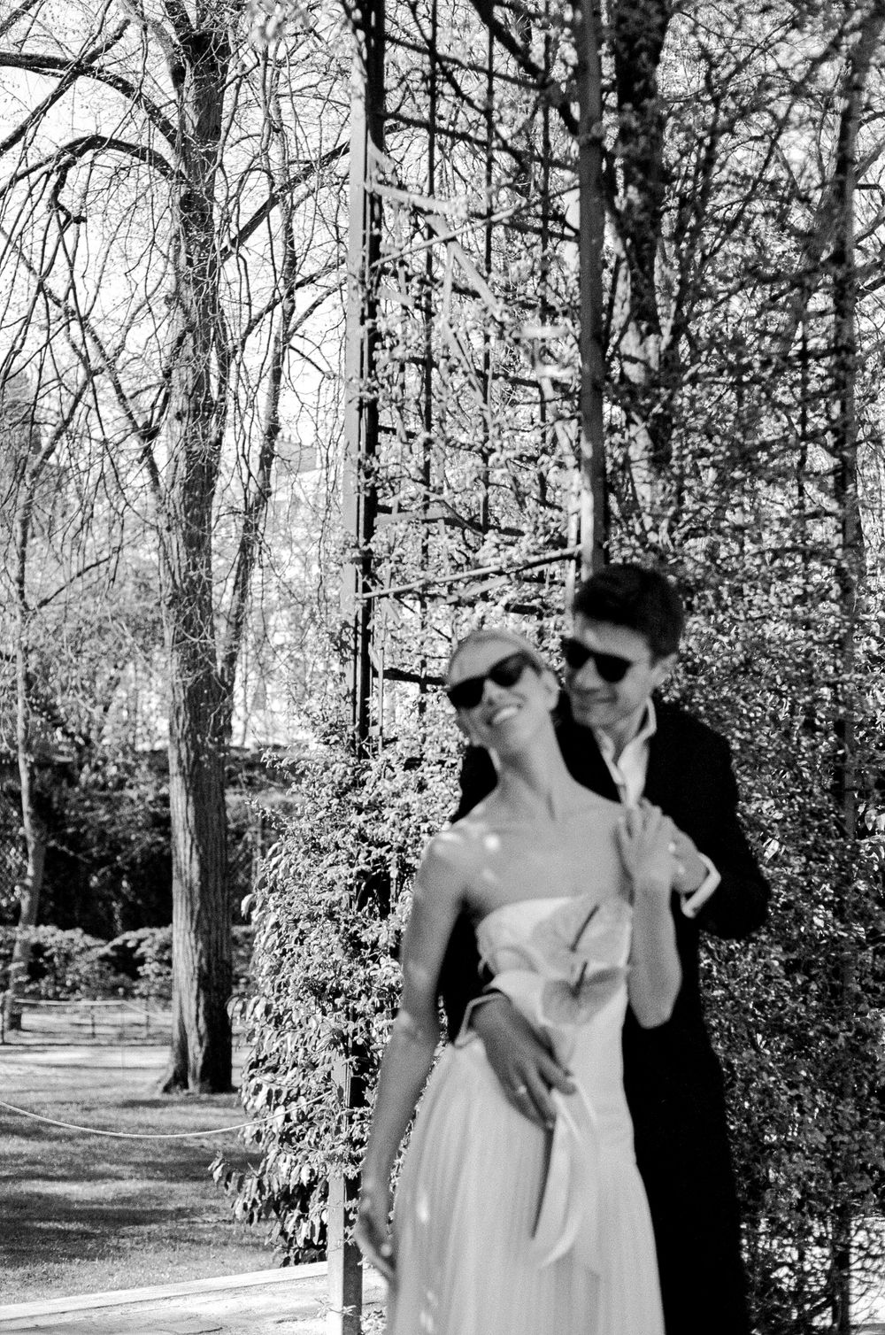 A black and white vintage photograph showing a romantic couple in formal attire embracing in a wooded garden setting.