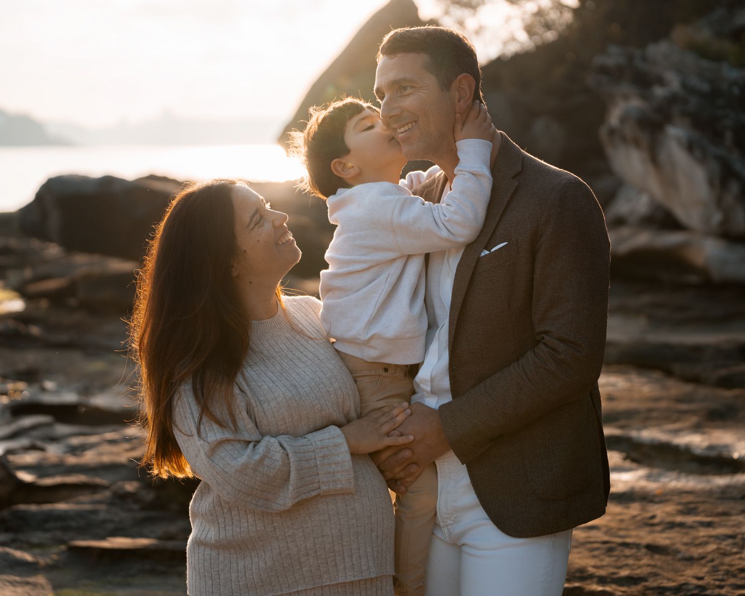 A young boy kisses his dad while his pregnant mom hugs them during a family photoshoot at a Sydney beach.