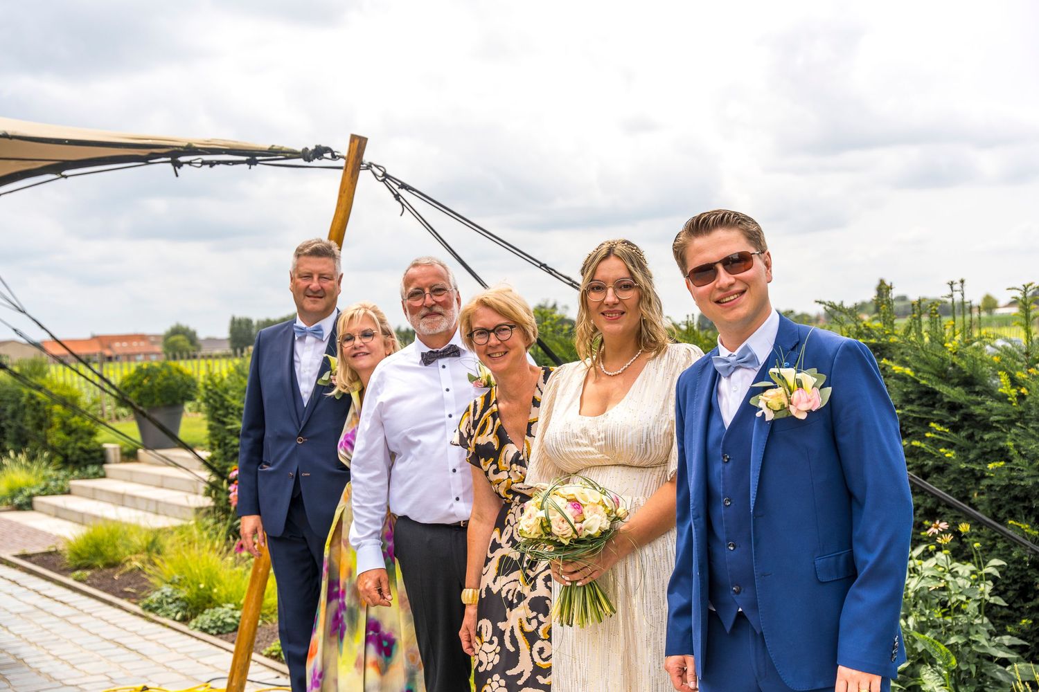 Wedding party gathered on outdoor terrace for group photo under cloudy sky.