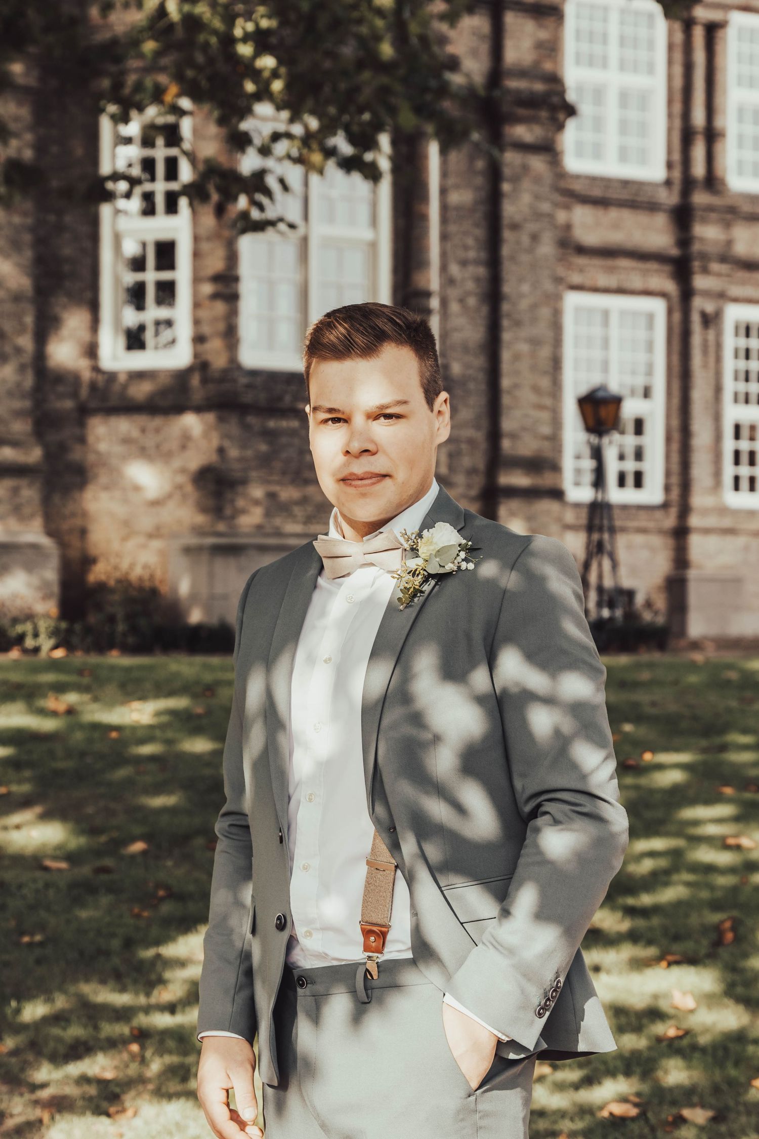 A groom in a grey suit and bow tie poses formally in front of a historic stone building.