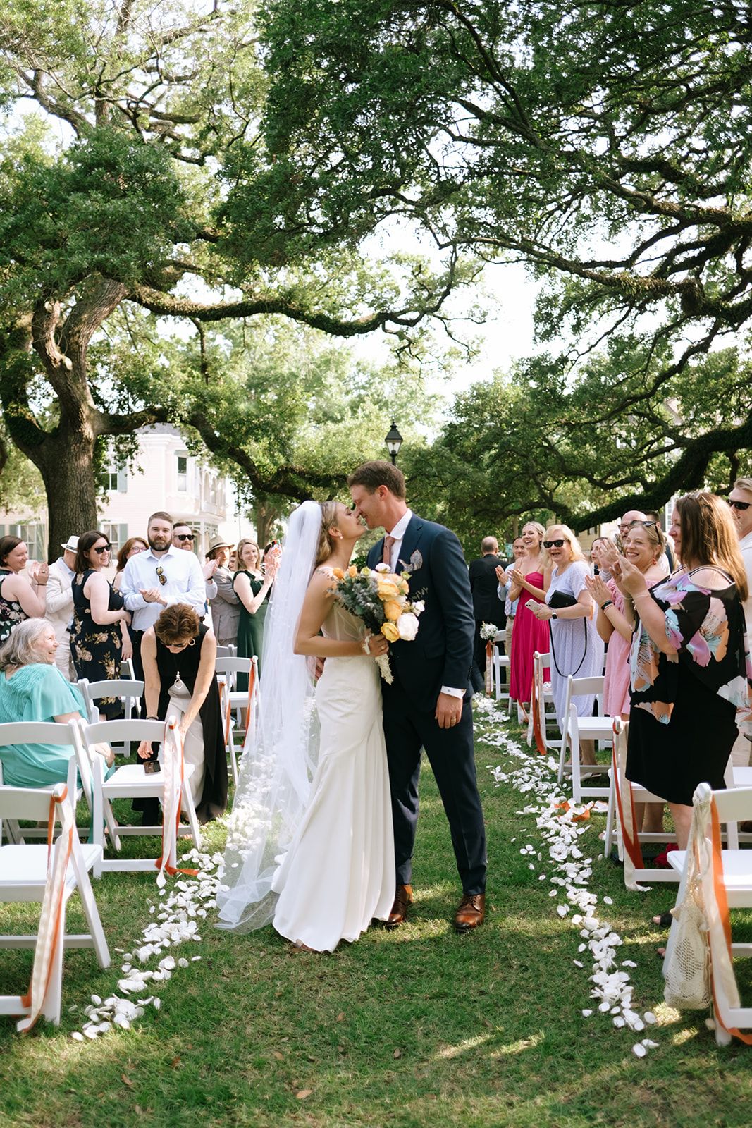 Bride and groom sharing first kiss at outdoor wedding ceremony under oak trees with guests.