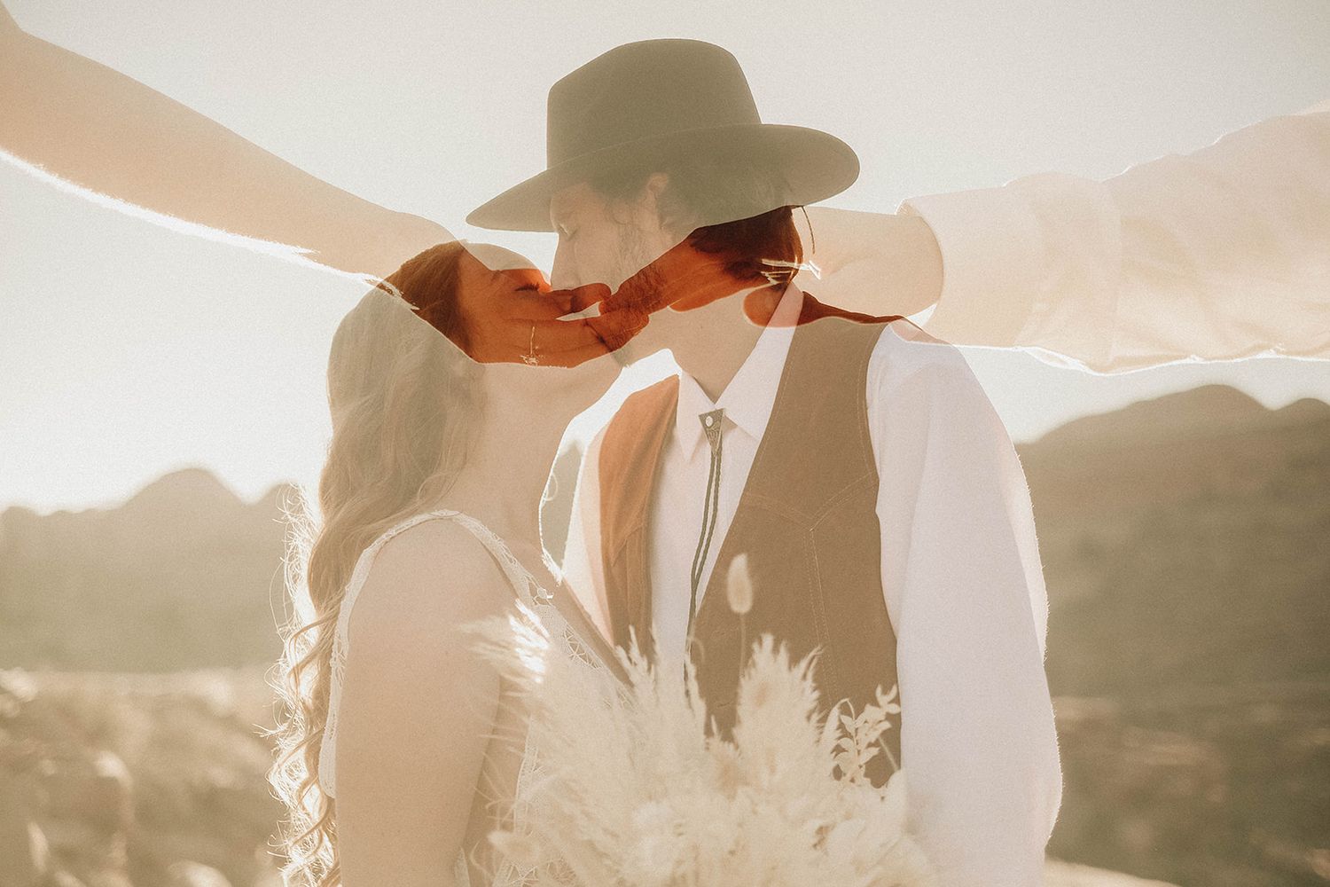 Bride and groom kissing in golden sunlight, groom wearing wide-brim hat and vest, desert backdrop.