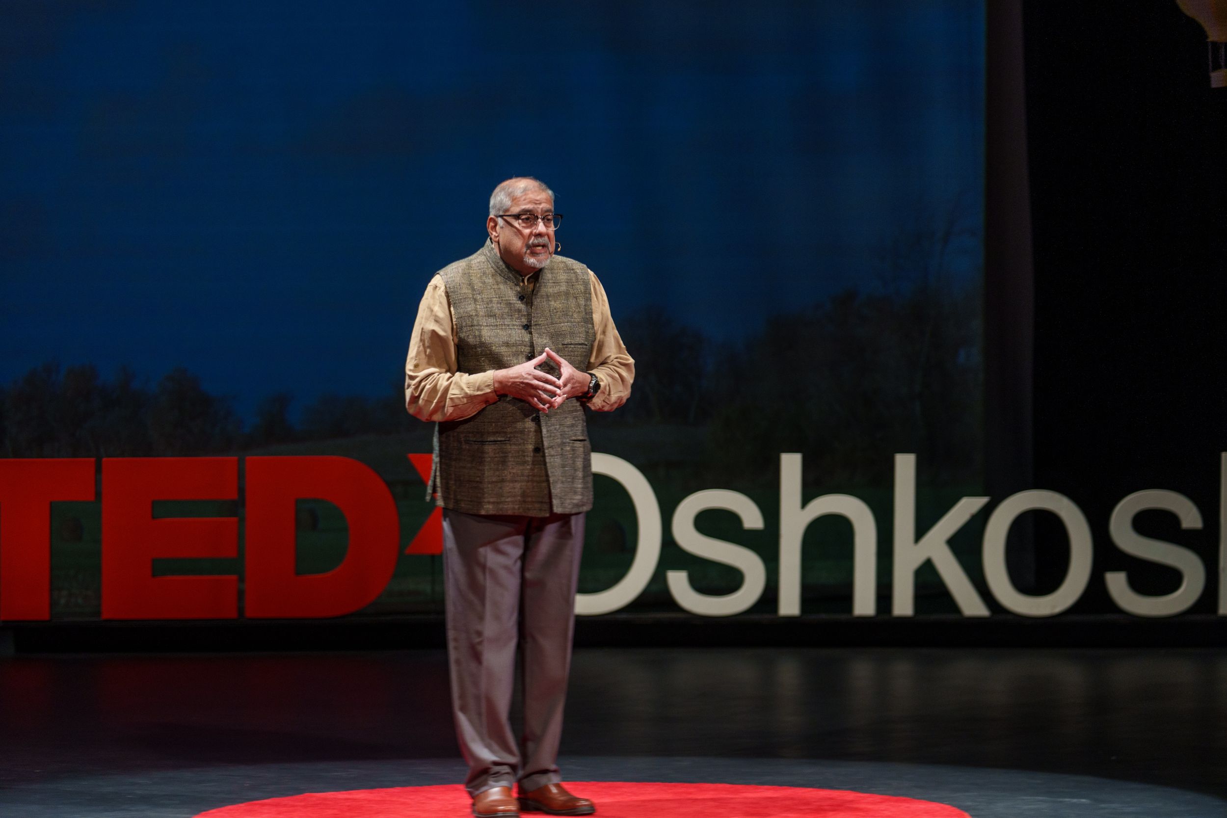 Speaker presenting on stage at TEDx Oshkosh event with red and white logo visible in background.