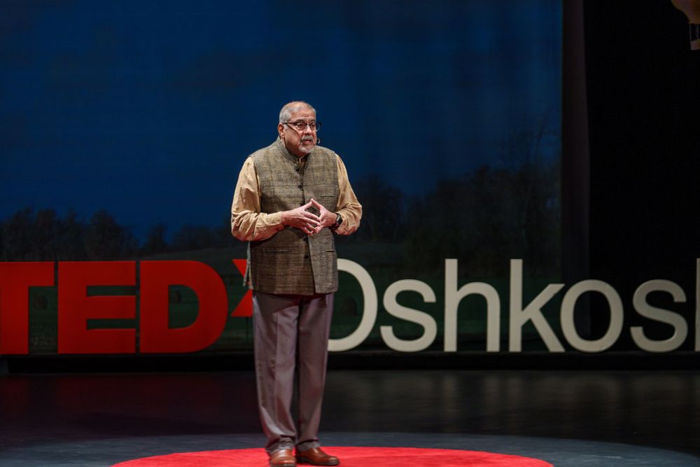 Speaker presenting on stage at TEDx Oshkosh event with red and white logo visible in background.