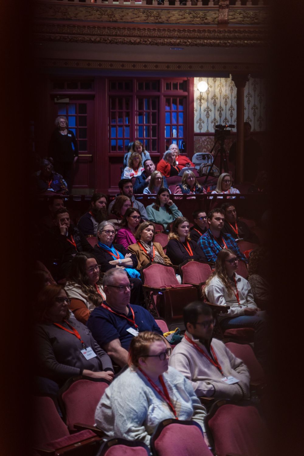 Audience members seated in red theater chairs watching a presentation in a dimly lit venue.