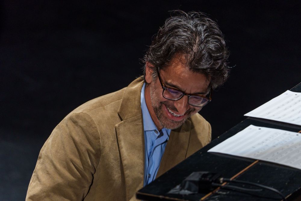 A pianist in a tan jacket smiles while performing at a grand piano on stage.