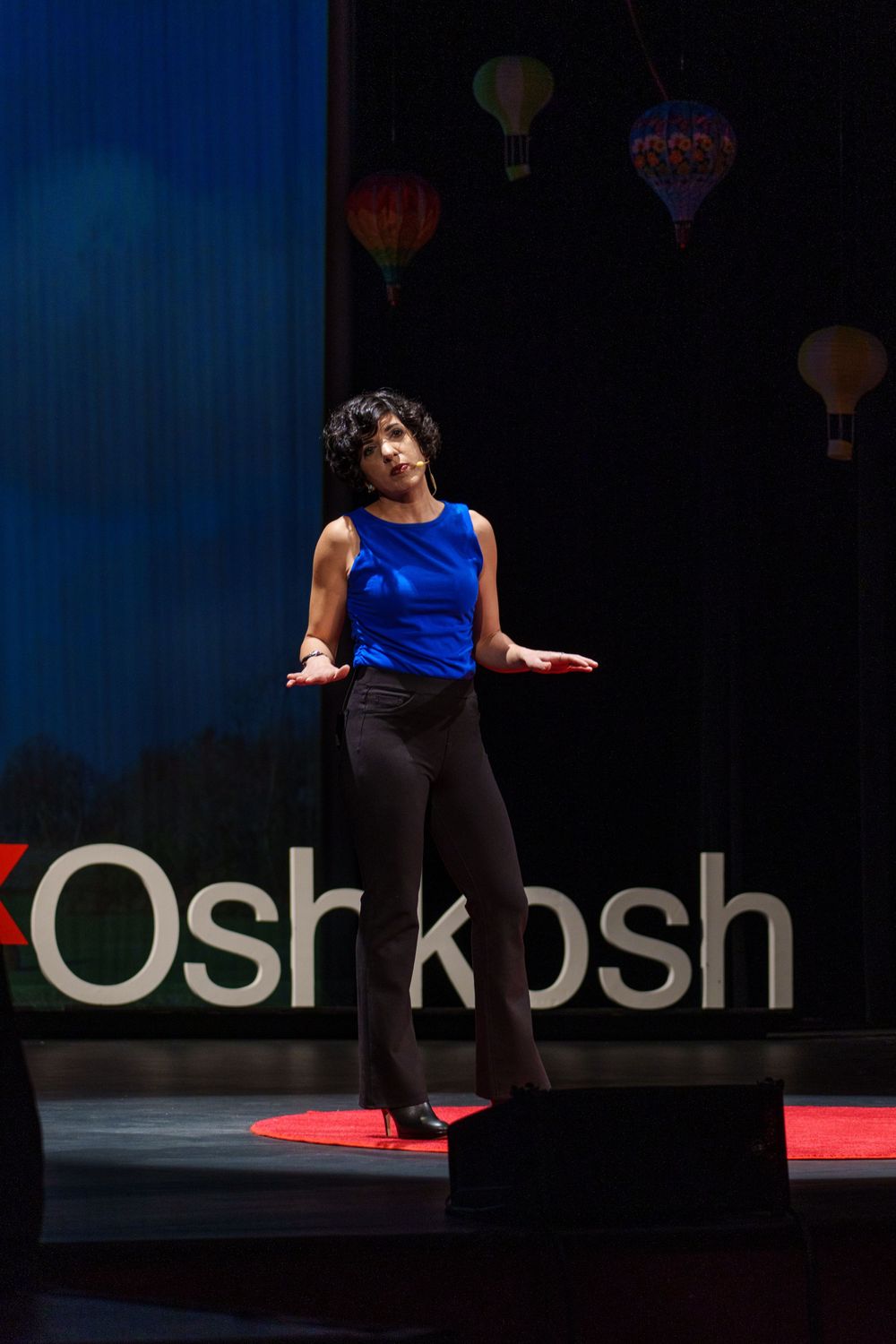 A presenter in blue speaks on the TEDx Oshkosh stage with balloon decorations in the background.