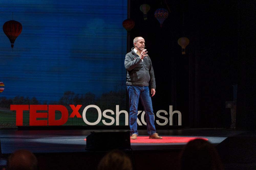 A speaker presents on stage at TEDx Oshkosh with red lighting and balloon decorations.