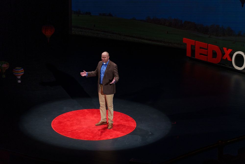A speaker stands in a spotlight on the red circular TEDx stage during a presentation.