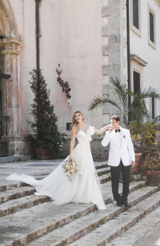Bride and groom posing on stone steps with tropical plants and architecture in background.