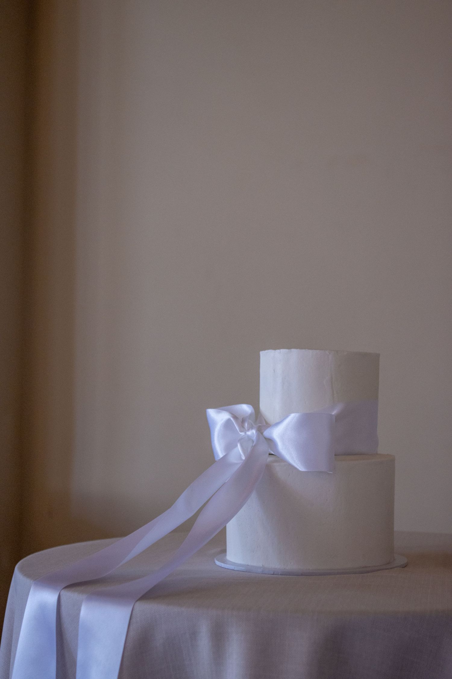 A simple two-tier white wedding cake decorated with a large satin ribbon bow on a round table.