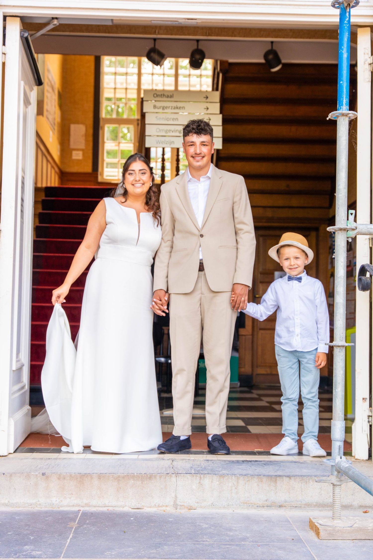 A wedding party stands in a building entrance with a red carpet and stairs behind them.