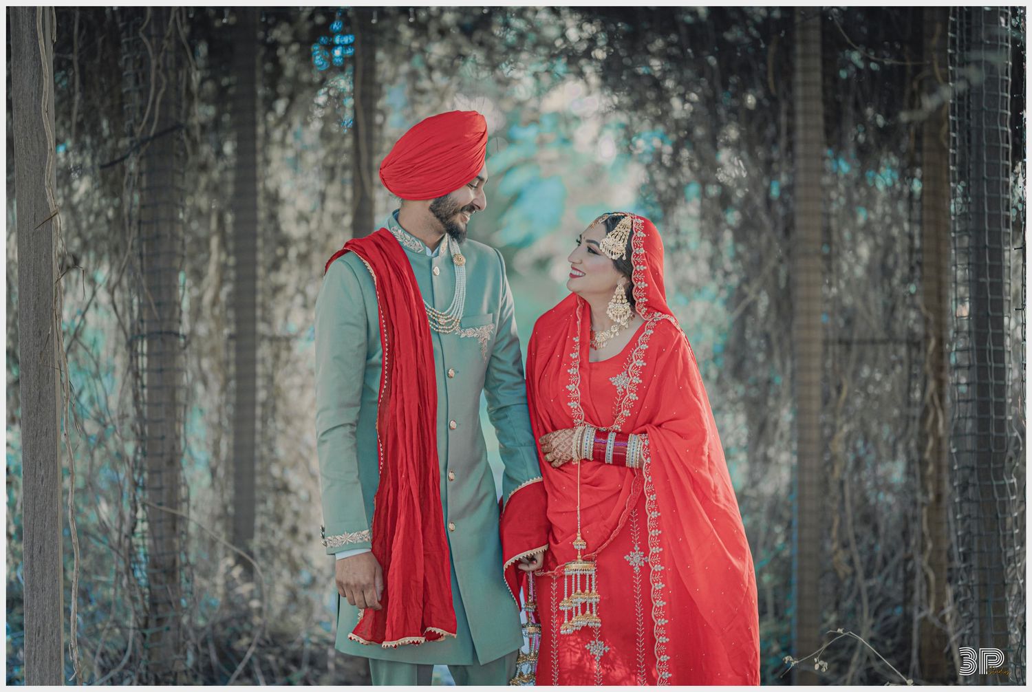 Couple in red traditional Punjabi wedding attire pose against ethereal forest backdrop.