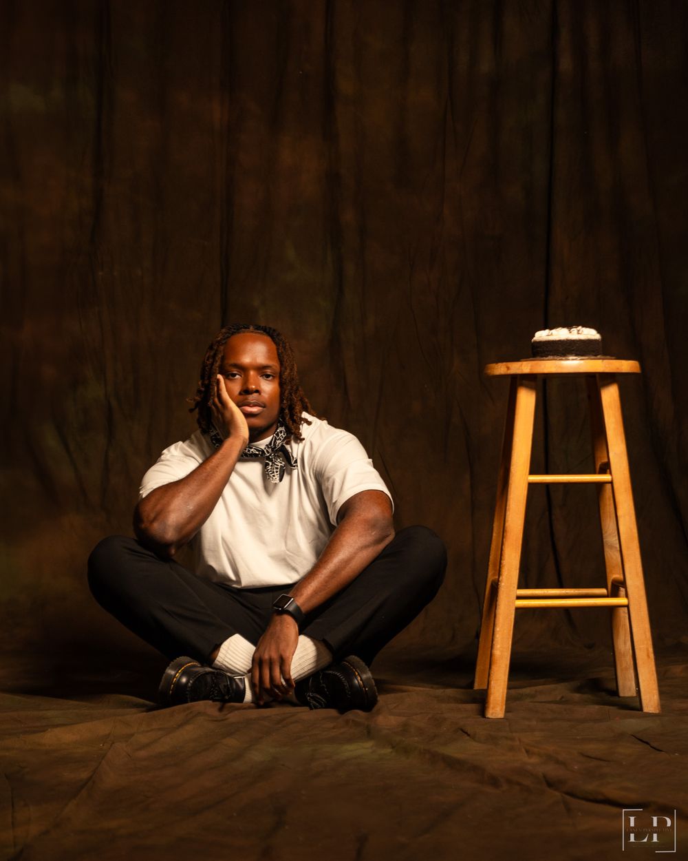 Person in white shirt and dark pants sits on floor near wooden stool in studio setting.