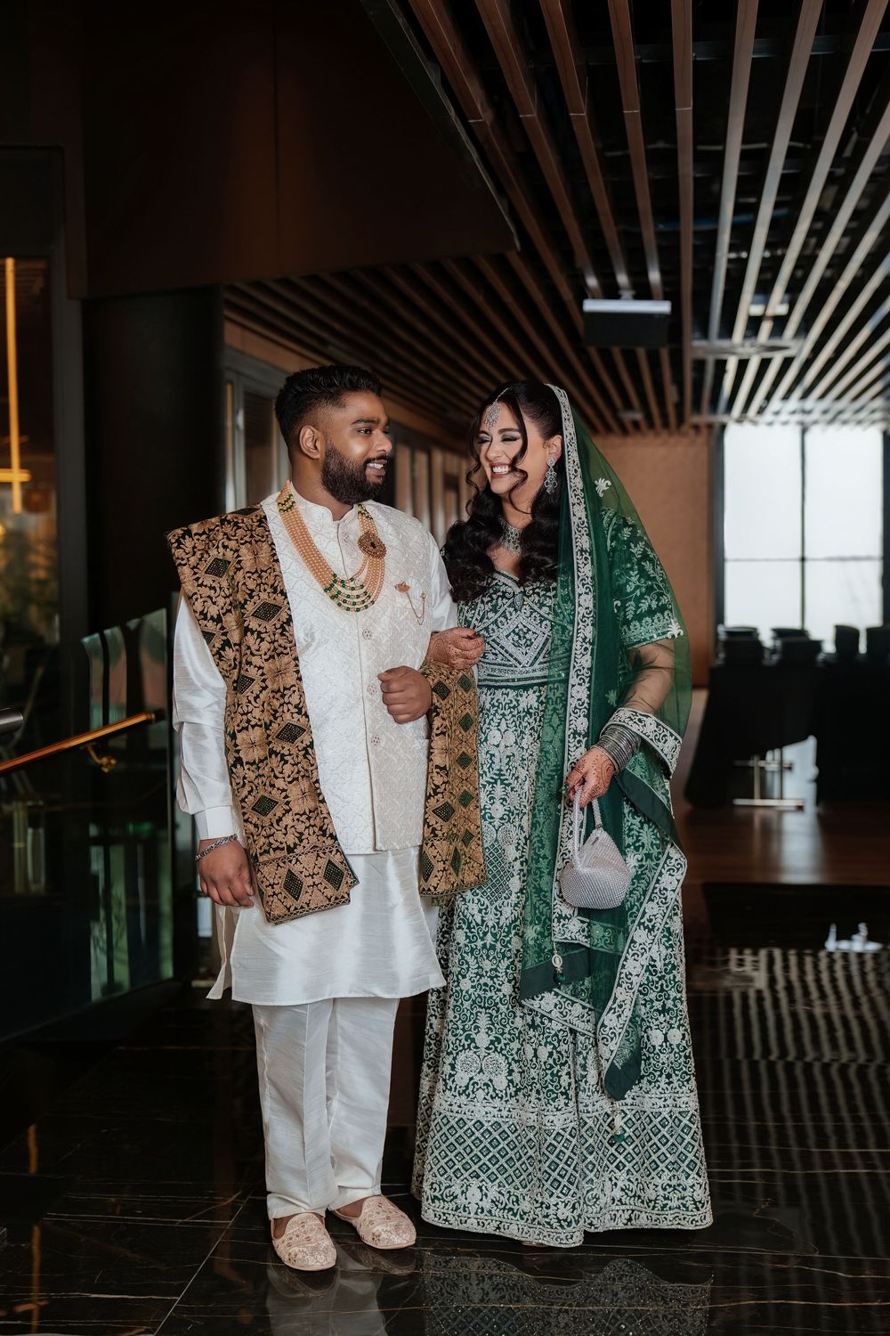 A couple in traditional South Asian attire, with a white kurta and gold vest paired with an emerald green embellished dress.