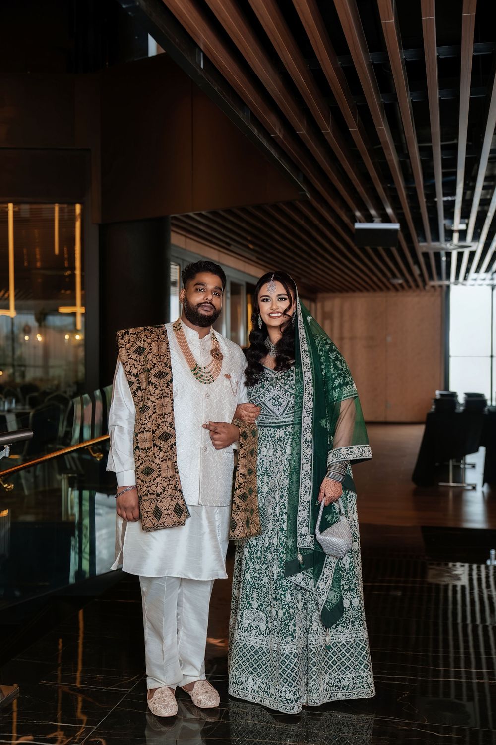 A couple in traditional South Asian wedding attire poses together, wearing ornate green and gold formal ethnic clothing.