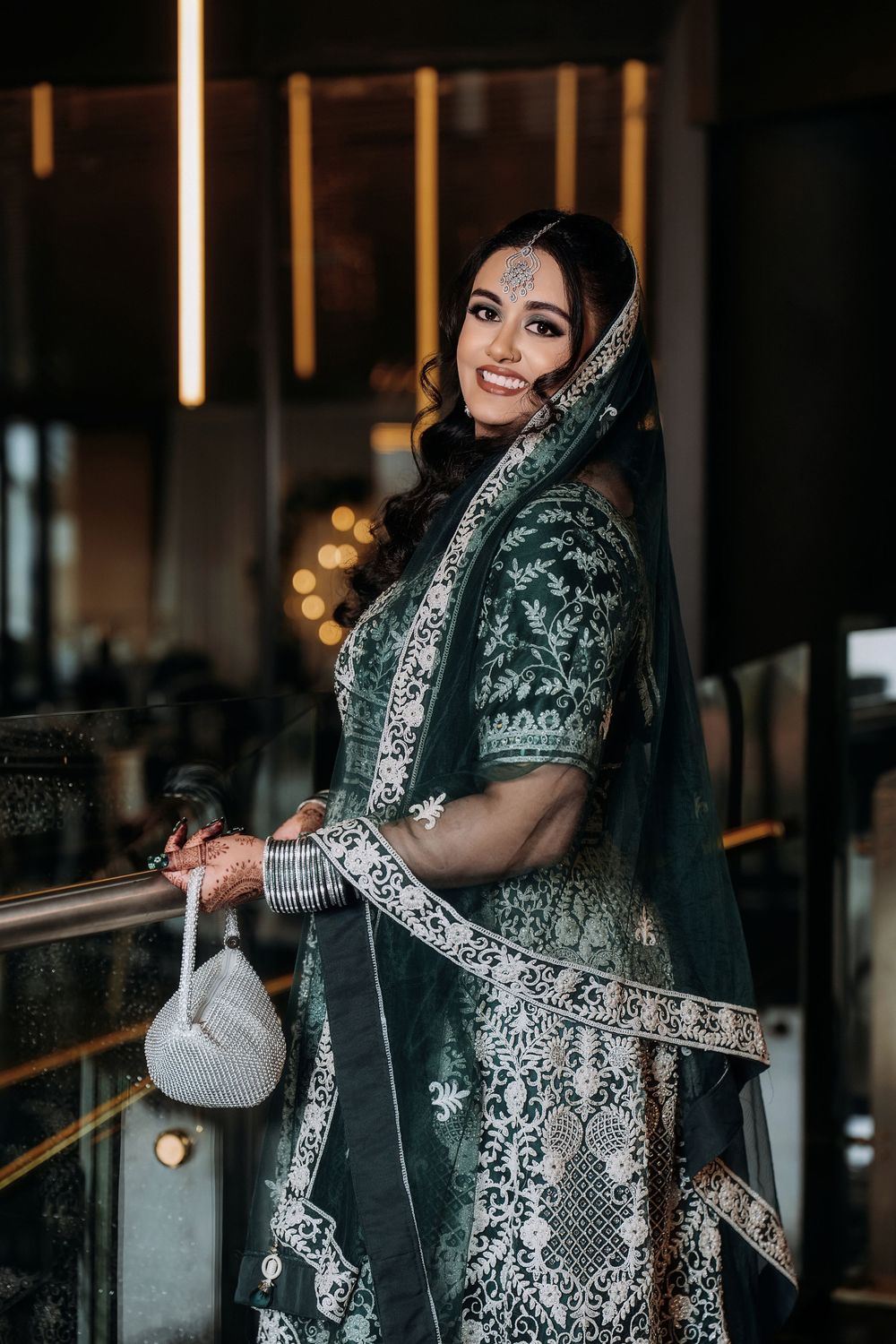 A woman in an elegant black and silver embroidered sari poses gracefully against a dark interior backdrop.