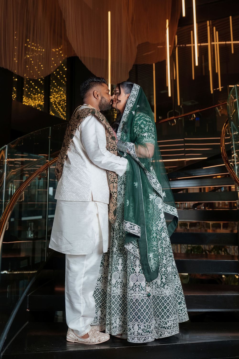 A couple in traditional wedding attire share an intimate moment on stairs, with the bride wearing an emerald green gown.