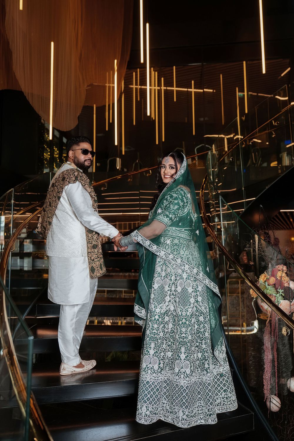 A couple in traditional South Asian wedding attire pose on an elegant staircase with glowing vertical lights in the background.