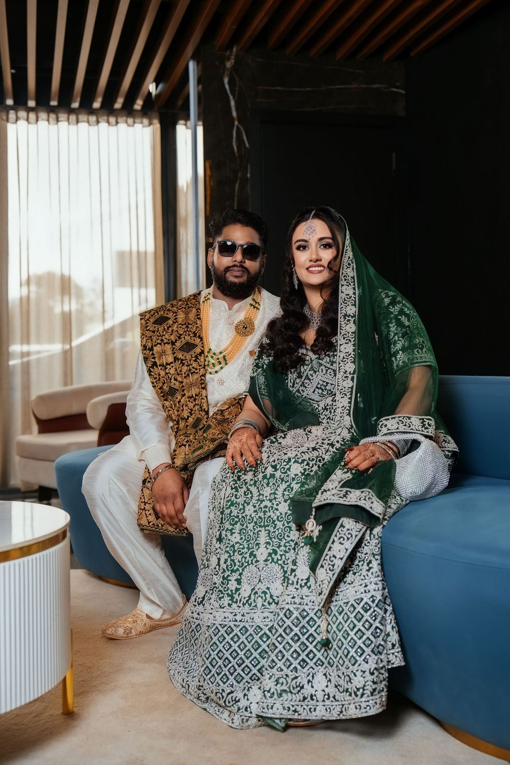 A couple in traditional Indian attire sits together on a blue couch in a modern interior setting.