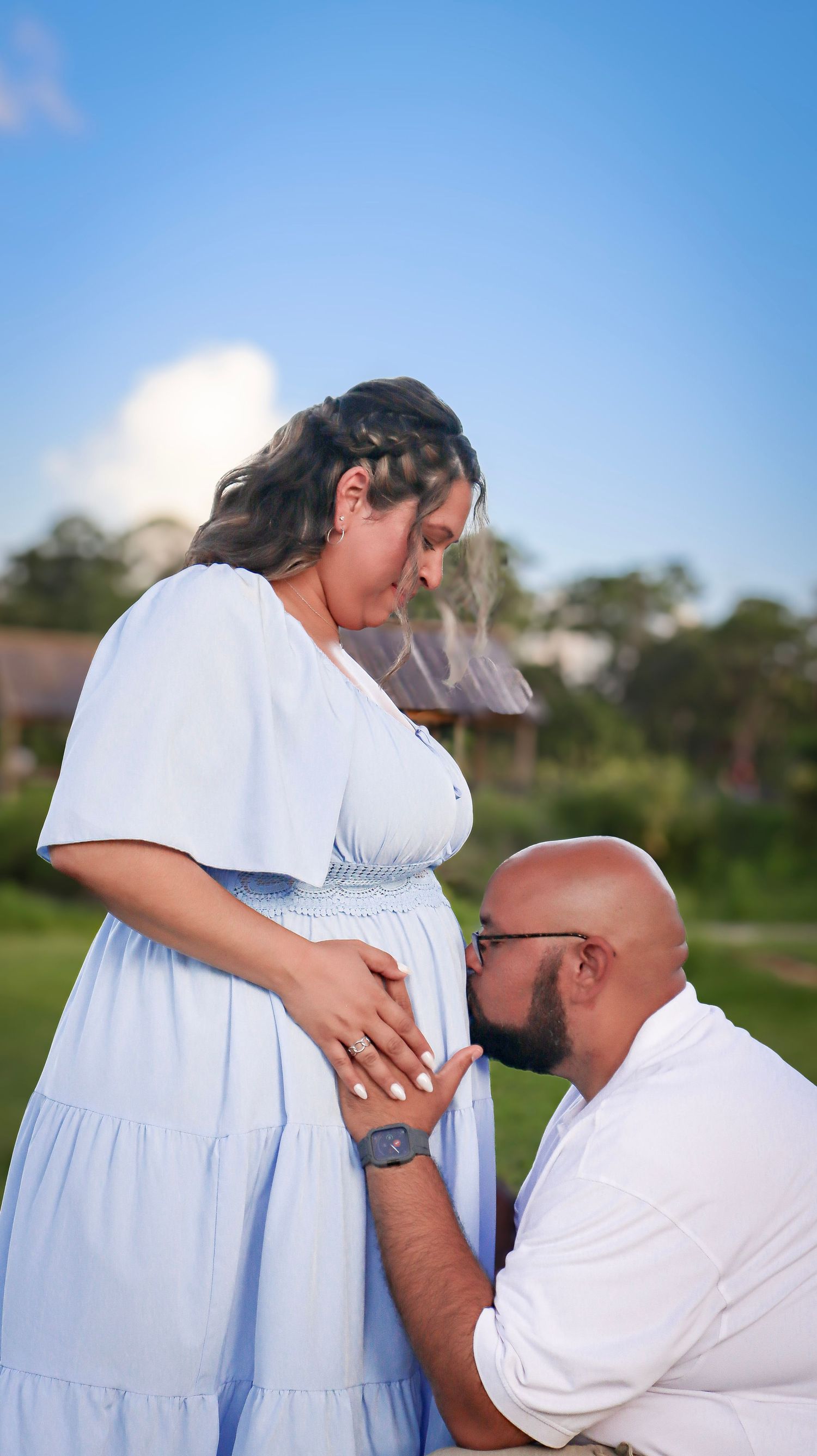 Couple shares tender moment outdoors in white outfits against blue sky background.