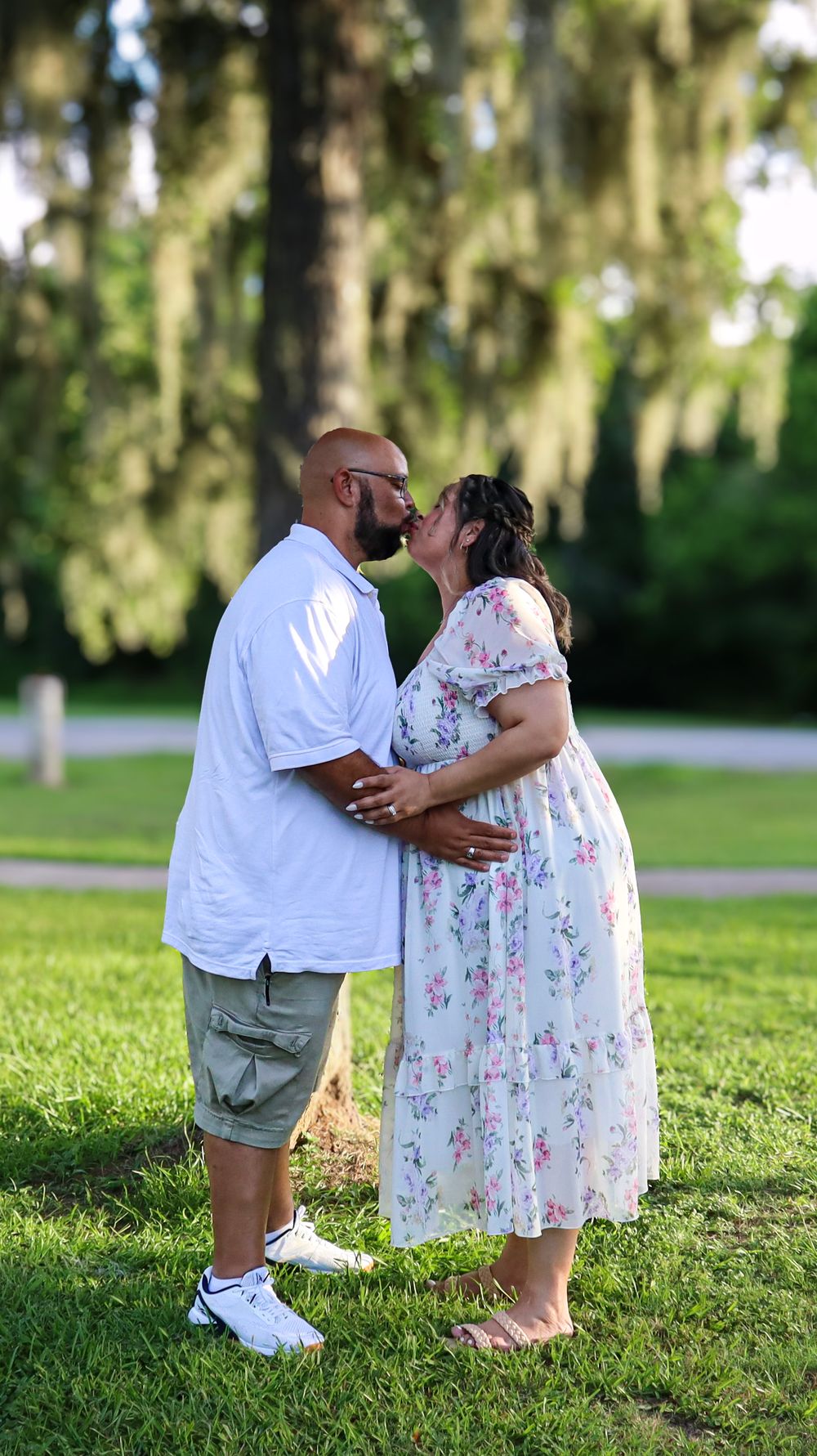 Couple shares romantic moment on grassy field with Spanish moss trees in background.