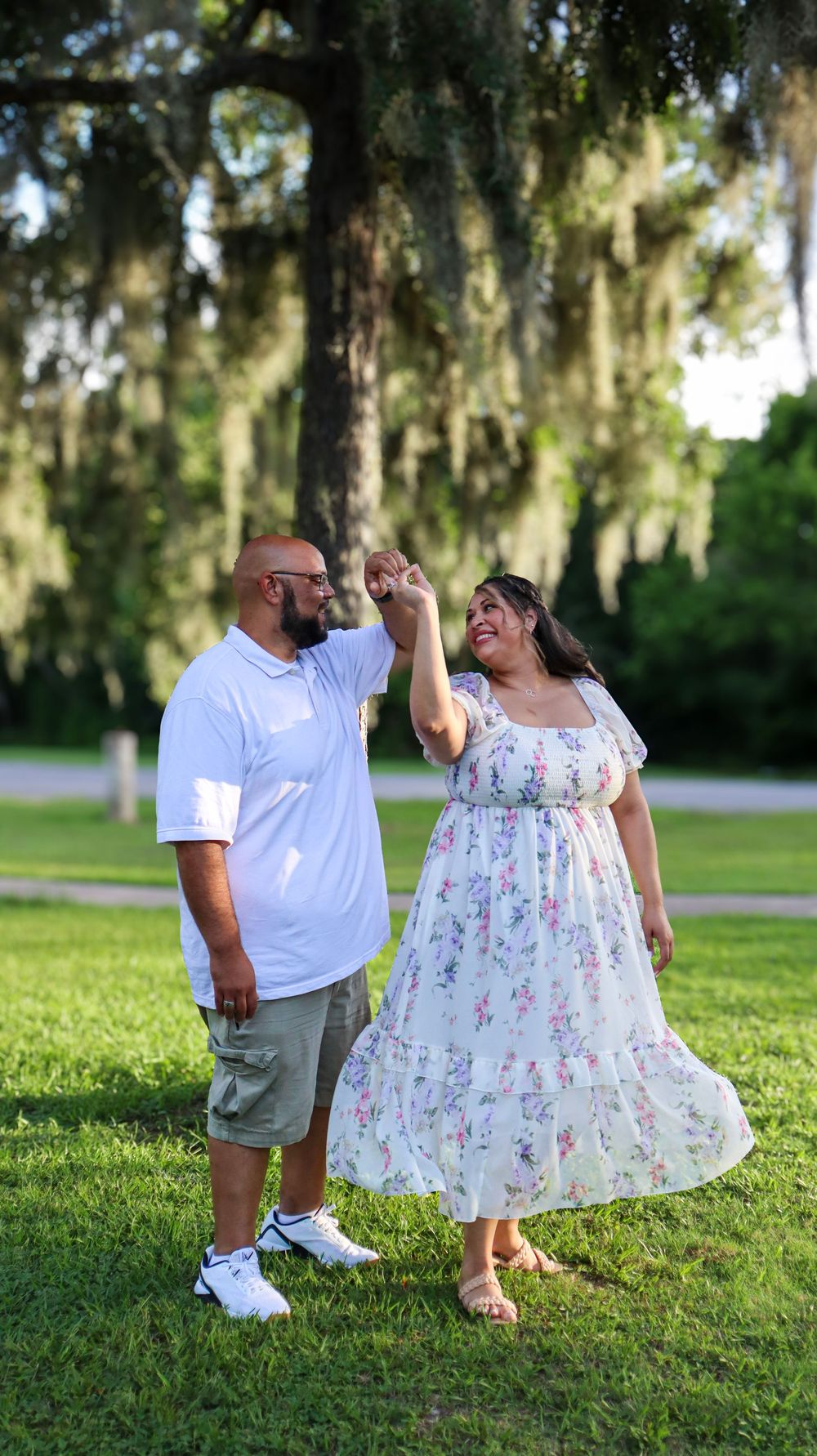 A couple holds hands while dancing and smiling in a park with moss-draped trees.
